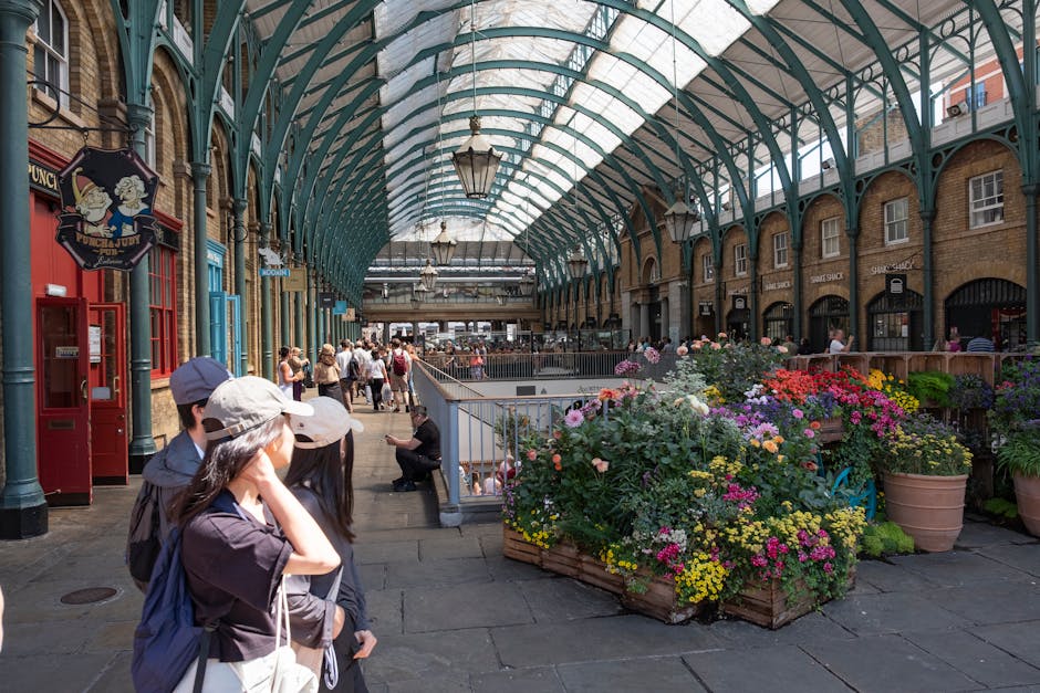 Inside a covered shopping arcade with an arched glass roof supported by green metal framework, a group of people, including a woman wearing a beige cap and a man with a baseball cap, are gathered on the pavement near an entrance. To the right, there are large wooden planters filled with colorful flowering plants, including pink, purple, yellow, and white blooms. The scene shows the surrounding brick and stone buildings with shopfronts and signage, and lantern-style lighting fixtures hanging from the ceiling. In the background, more shoppers are walking and browsing, with some seated on benches. A woman in black attire is sitting on the steps, and nearby, a man is using his mobile phone. This setting depicts a lively indoor marketplace or shopping arcade, with natural daylight filtering through the glass roof, all within a vibrant, historic urban environment. Occasionally, Man with Van Covent Garden's presence in house removals and furniture transport processes is subtly supported by the scene's focus on logistical movement and pedestrian activity.