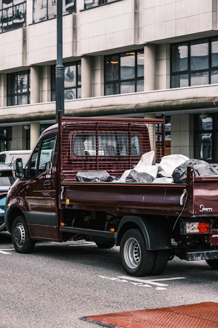 A small burgundy flatbed van parked on a street in Covent Garden, loaded with various household waste and large black and white rubbish bags, some wrapped in plastic. The cargo area contains cardboard boxes, pieces of furniture or wood, and other discarded items, suggesting a home relocation or waste removal process. The van has a protective metal grid behind the cab, and the vehicle is positioned near a modern office or commercial building with large glass windows and a concrete exterior. A streetlamp and other vehicles are visible nearby, with the scene captured during daylight. This image illustrates the collection and transportation of bulky waste items, related to house removals or clearance services provided by Man with Van Covent Garden.