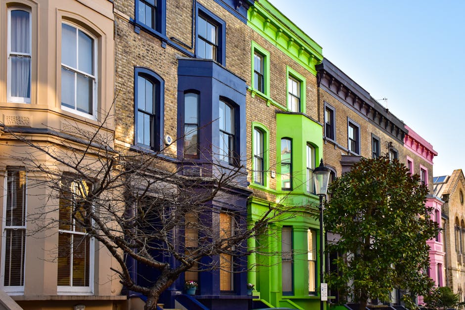 A row of multi-storey Victorian terraced houses in Covent Garden, London, painted in bright, distinct colours including cream, dark blue, lime green, and pink. The buildings feature large sash windows and decorative stonework. A leafless tree with thin branches partially obscures the lower portion of the houses, standing on the pavement in front of the buildings. A traditional black street lamp is positioned near the lime green house, which is adjacent to the cream and dark blue buildings, with similar street fixtures nearby. The scene is illuminated by natural daylight, highlighting the vibrant hues of the façades. This image depicts a residential area often associated with urban home relocation and furniture transport, illustrating the environment where professional removals services such as those offered by Man with Van Covent Garden are frequently required during house moves or packing and moving tasks inside the city.