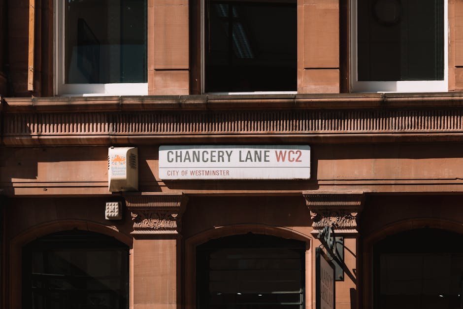 Photograph of a building facade on Chancery Lane in Westminster with a street sign indicating WC2. The sign is white with black and red text, mounted above an arched entranceway. The building is constructed from reddish-brown stone or brick with detailed architectural elements around the windows and doorways. To the left of the street sign, there's a small external box, possibly an electrical or communication box. The image is taken in natural daylight, highlighting the texture and colors of the stonework. This scene may be part of a home relocation process or furniture transport setup near a property on Chancery Lane, where professional removals such as those by Man with Van Covent Garden could facilitate packing, loading, and transportation of household items across the local area.