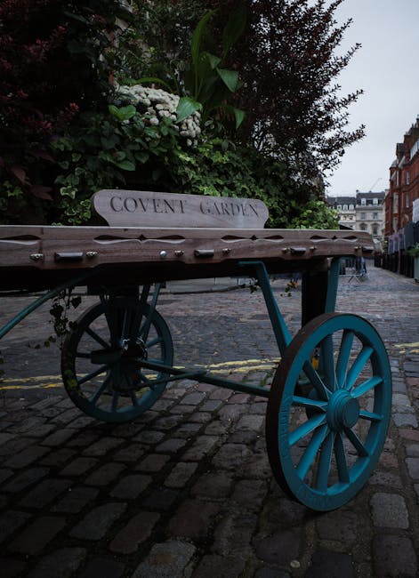 A wooden handcart with blue-painted wheels and a flat wooden surface is positioned on a cobblestone pavement outside a residential area, near a garden hedge with flowering plants and greenery. The cart has a white sign attached to its side with the text 'COVENT GARDEN' engraved on it. In the background, there are multi-storey buildings lining the street under an overcast sky. The scene appears to be set during daytime, with soft natural light. The cart is part of a home relocation or furniture transport setup, and the setting suggests a street-side loading area for a house removal service, which might be handled by [COMPANY_NAME]. The environment includes typical London-style architecture, and the conveyor features tools and materials for moving household furniture and boxes, supporting the context of packing and loading during a house move.