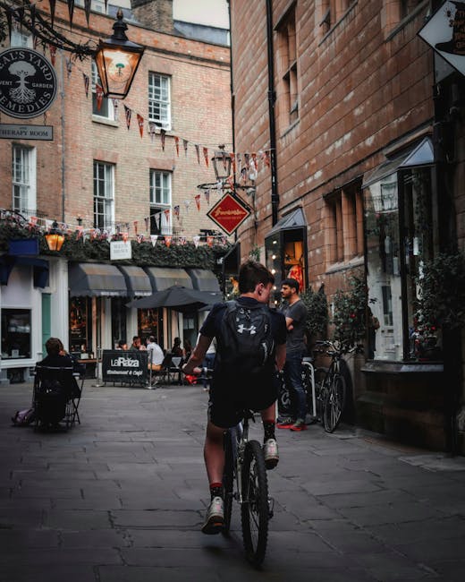 A person riding a bicycle along a narrow pedestrian street in Covent Garden, London, during daytime. The cyclist is wearing a dark backpack and sports shorts, and is approaching a small group of peopl