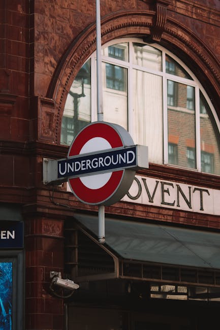 A close-up view of the London Underground sign hanging outside a historic red-brick building with large arched windows, situated in Covent Garden. In the foreground, the sign displays the word 'UNDERG