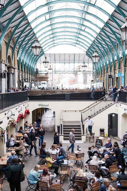 Interior view of a busy indoor market with a high, curved glass ceiling supported by teal metal arches. The space features multiple levels, with a balcony overlooking a ground floor filled with seated patrons at wooden tables and chairs. People are engaged in conversations, dining, and walking through the area. Stairs lead up to the balcony, which has black railings and is lined with several standing individuals. Modern lantern-style lighting fixtures hang from the ceiling, and the market is illuminated by natural light coming through the large glass roof. The environment suggests a vibrant yet orderly atmosphere typical of a market or food hall, suitable for home relocation or moving logistics, with visible elements like furniture, tables, and general interior features associated with a busy commercial space managed by a professional removals service like Man with Van Covent Garden.