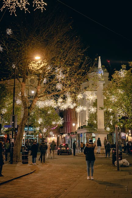 A nighttime street scene near Seven Dials in Covent Garden with festive holiday lights illuminating trees and shopfronts. In the foreground, several pedestrians are walking along the cobbled pavement, some wearing coats and hats. To the right, a delivery van belonging to Man with Van Covent Garden is parked near the curb, partially loaded with furniture and packing boxes wrapped in plastic and cardboard. The van's rear doors are open, and a few items, including a large wooden piece and stacked boxes, are being loaded or unloaded. The surrounding buildings have decorative façades and illuminated shop signs, with streetlights providing additional lighting. The environment suggests a home relocation or furniture transport process, with careful handling of household items in an active, urban area.