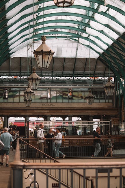 Interior view of a busy indoor market with a high, curved glass ceiling supported by teal metal arches. The space features multiple levels, with a balcony overlooking a ground floor filled with seated patrons at wooden tables and chairs. People are engaged in conversations, dining, and walking through the area. Stairs lead up to the balcony, which has black railings and is lined with several standing individuals. Modern lantern-style lighting fixtures hang from the ceiling, and the market is illuminated by natural light coming through the large glass roof. The environment suggests a vibrant yet orderly atmosphere typical of a market or food hall, suitable for home relocation or moving logistics, with visible elements like furniture, tables, and general interior features associated with a busy commercial space managed by a professional removals service like Man with Van Covent Garden.