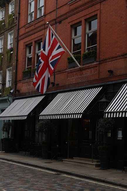 A red brick building with large windows and black-and-white striped awnings extends over a cobbled street pavement. A Union Jack flag is mounted on a pole attached to the building, fluttering slightly in the breeze. The ground floor appears to be an establishment, possibly a pub or restaurant, with outdoor seating and large planters positioned along the sidewalk. The scene is well-lit, indicating daytime, and suggests an urban setting suitable for house removals or furniture transport. The image captures an area that may be affected by street closures or parking regulations, relevant to relocation services such as those provided by Man with Van Covent Garden.