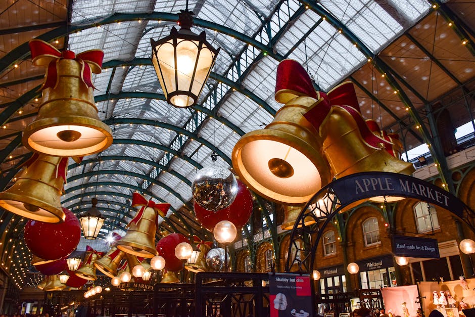Inside a covered market with a high, curved glass ceiling, large gold decorative bells with red ribbons, and silver and red spherical ornaments hanging from the ceiling. Several traditional lantern-style lights provide illumination. In the foreground, a black metal cart or trolley is positioned near a sign reading 'APPLE MARKET'. The environment appears festive, likely set up for a holiday event or seasonal display. The scene includes packaging materials such as cardboard boxes and plastic wrap, suggesting preparations related to home relocation or moving services. The presence of a hoisting or loading area indicates an active moving process, with potential for furniture transport or packing and moving activities, associated with house removals, as offered by Man with Van Covent Garden.